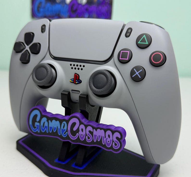 Puede incluir: Un mando gris de PlayStation 5 con botones y joysticks negros, apoyado en un soporte negro y morado. El soporte presenta el texto "Game Cosmos" en azul y morado. El logotipo de PlayStation es visible.