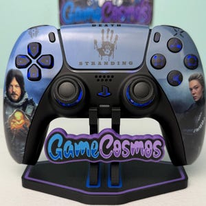 Può includere: Un controller PlayStation 5 personalizzato con opere d'arte del gioco Death Stranding. Il controller ha un design blu e nero con ritratti di personaggi e il logo del gioco. È esposto su un supporto con il logo "Game Cosmos".