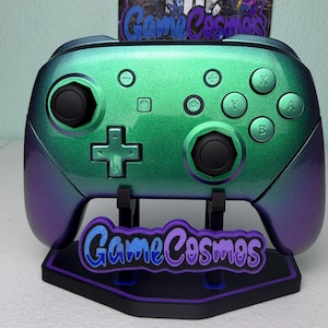 May include: A wireless video game controller with a metallic green and purple color-shifting finish, featuring black joysticks, a directional pad, and action buttons. It rests on a black stand with the text GameCosmos in blue and purple.
