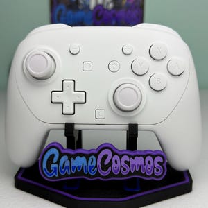 Può includere: Un controller di gioco bianco con pulsanti e joystick, appoggiato su un supporto. Il supporto è nero con un logo viola e blu che dice "Game Cosmos". Il controller ha un D-pad, pulsanti funzione e pulsanti dorsali.