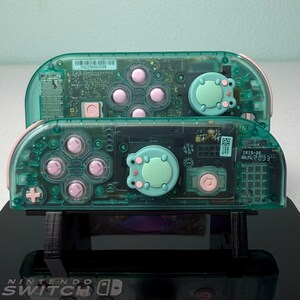 May include: Two transparent green Nintendo Switch Joy-Cons with pink buttons and a light green frog-shaped button on each. The Joy-Cons are on a black stand with the Nintendo Switch logo.