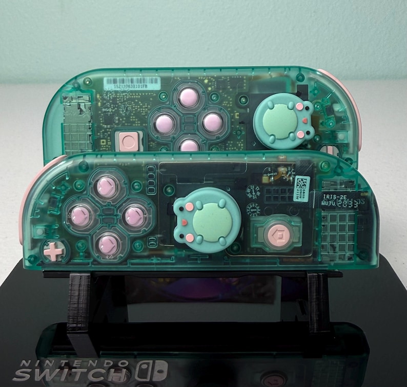 May include: Two transparent green Nintendo Switch Joy-Cons with pink buttons and a light green frog-shaped button on each. The Joy-Cons are on a black stand with the Nintendo Switch logo.