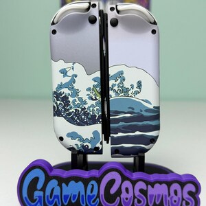 May include: A pair of white and gray game controllers with a wave design, mounted on a stand with the text "Game Cosmos" in blue and purple. The controllers are designed for a gaming console.