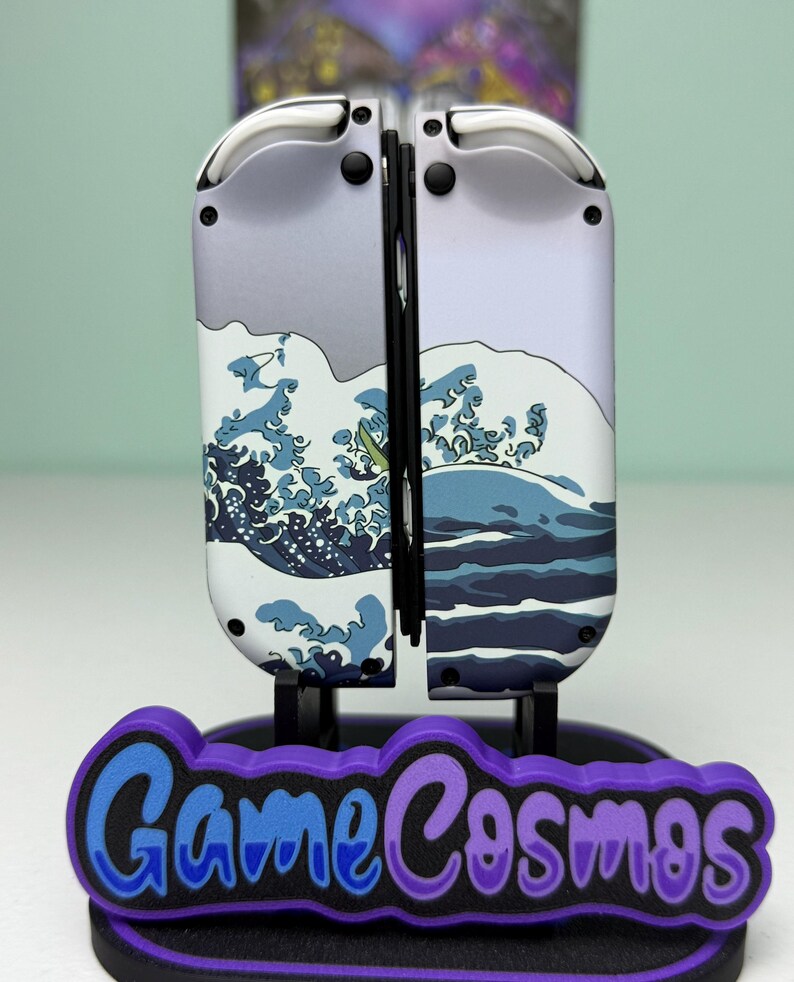 May include: A pair of white and gray game controllers with a wave design, mounted on a stand with the text "Game Cosmos" in blue and purple. The controllers are designed for a gaming console.