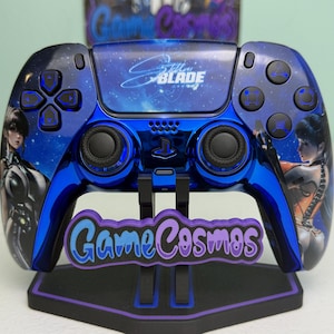 以下が含まれることがあります： 「Stellar Blade」デザインのカスタムブルーとブラックのPlayStation 5コントローラー。女性のアートワークと「Game Cosmos」のロゴがスタンドに表示されています。