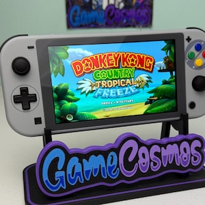 May include: A gray handheld gaming console displaying the "Donkey Kong Country: Tropical Freeze" game. The console is on a black stand with the text "Game Cosmos" in purple and blue. The console has a black D-pad and buttons.