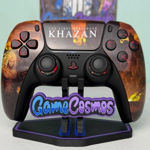 May include: A black wireless game controller featuring a custom orange and brown graphic wrap with the text The First Berserker KHAZAN. The controller has red accents on the buttons and joysticks, resting on a black stand with a GameCosmos logo.