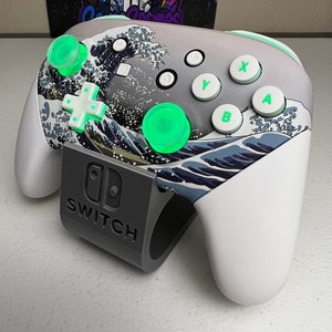 Puede incluir: Un mando blanco de Nintendo Switch con un dise&ntilde;o de la Gran Ola de Kanagawa. El mando tiene botones verdes y una base gris con la palabra "SWITCH" impresa.