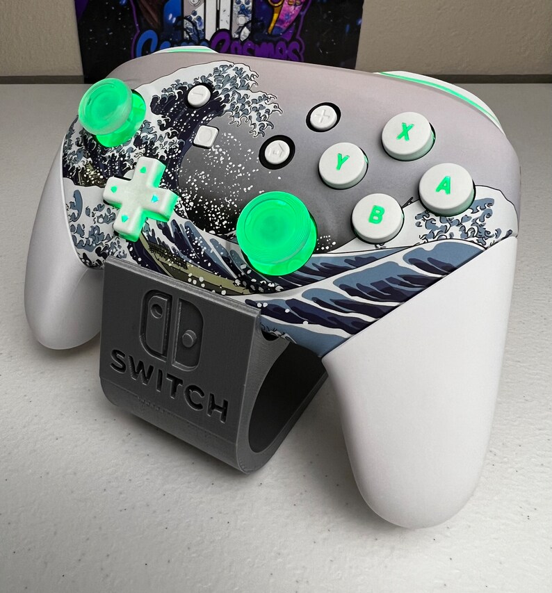 Puede incluir: Un mando blanco de Nintendo Switch con un dise&ntilde;o de la Gran Ola de Kanagawa. El mando tiene botones verdes y una base gris con la palabra "SWITCH" impresa.