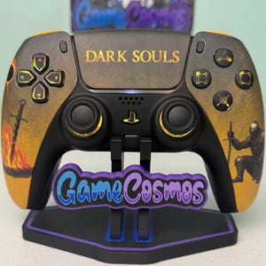 May include: A custom black and gold PlayStation 5 controller with "DARK SOULS" text. The controller features artwork of a knight and a burning sword. The controller is on a stand with "Game Cosmos" text.