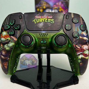 Könnte beinhalten: Ein grüner und schwarzer PlayStation 5-Controller mit Teenage Mutant Ninja Turtles-Grafiken. Der Controller hat eine grüne, durchsichtige Hülle und schwarze Tasten. Die Vorderseite des Controllers zeigt ein Gullydeckel-Design mit den Worten "Teenage Mutant Ninja Turtles" und "Turtles" darauf gedruckt. Die Rückseite des Controllers zeigt Bilder der Teenage Mutant Ninja Turtles.