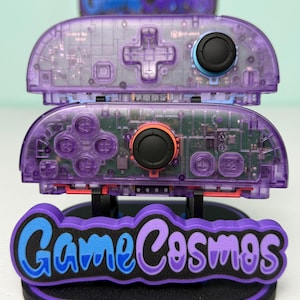 Può includere: Controller di gioco trasparenti viola impilati su un supporto nero con il testo "Game Cosmos" in blu e viola. I controller hanno joystick e pulsanti neri. Il supporto è su una superficie verde chiaro.