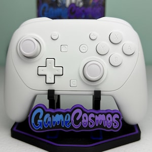 May include: A white wireless video game controller featuring two analog sticks, a directional pad, and action buttons labeled X, Y, A, and B. The controller rests on a black stand with a purple and blue GameCosmos logo plate attached.