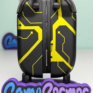 Puede incluir: Dispositivo electr&oacute;nico negro y amarillo con dise&ntilde;o de placa de circuito. El dispositivo est&aacute; sobre una base morada y azul con el texto "Game Cosmos". El dispositivo tiene un dise&ntilde;o con bisagras.