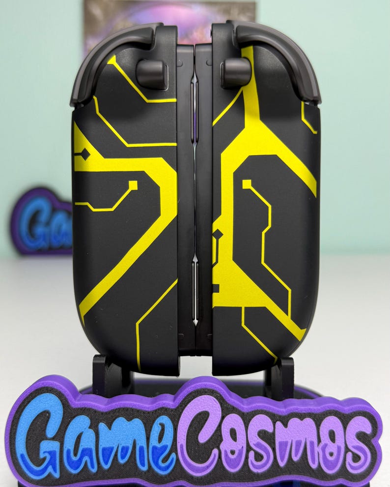 Puede incluir: Dispositivo electr&oacute;nico negro y amarillo con dise&ntilde;o de placa de circuito. El dispositivo est&aacute; sobre una base morada y azul con el texto "Game Cosmos". El dispositivo tiene un dise&ntilde;o con bisagras.