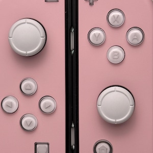 Może przedstawiać: R&oacute;żowa konsola Nintendo Switch z białymi przyciskami i czarnym zawiasem. Konsola posiada krzyżak, cztery przyciski, dwa przyciski na ramionach i przycisk home.