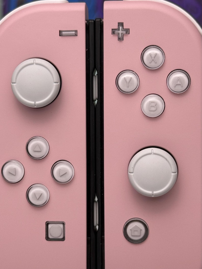 Może przedstawiać: R&oacute;żowa konsola Nintendo Switch z białymi przyciskami i czarnym zawiasem. Konsola posiada krzyżak, cztery przyciski, dwa przyciski na ramionach i przycisk home.