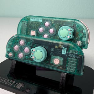 May include: Two teal green Nintendo Switch Joy-Con controllers with pink buttons and custom green and pink cat-shaped button covers. The controllers are on a black stand with the Nintendo Switch logo.