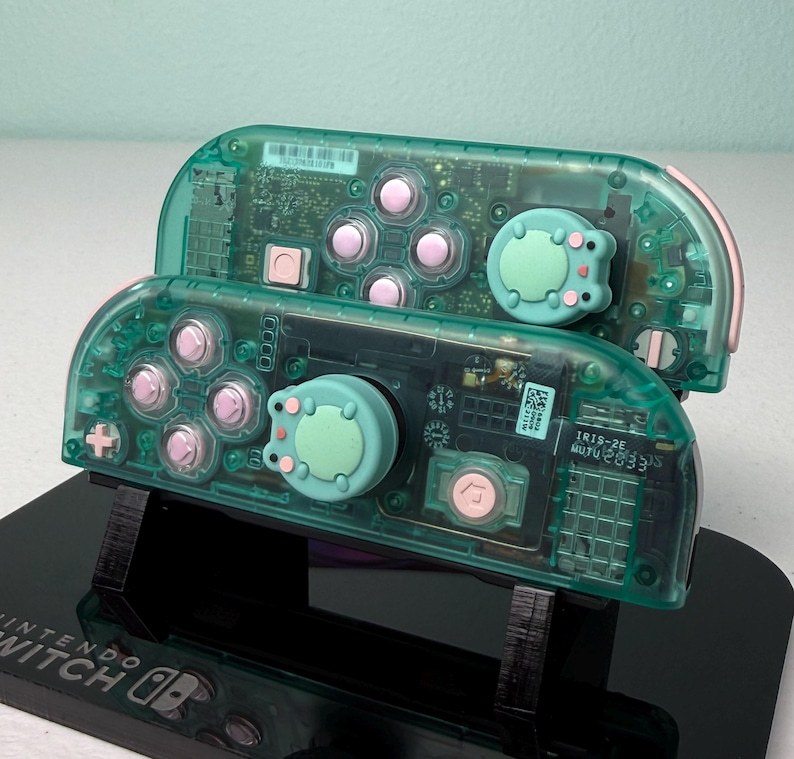 May include: Two teal green Nintendo Switch Joy-Con controllers with pink buttons and custom green and pink cat-shaped button covers. The controllers are on a black stand with the Nintendo Switch logo.