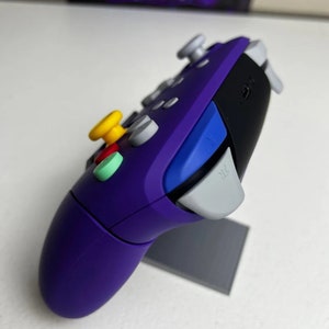 May include: Purple and black video game controller with a yellow, red, and green button. The controller has a gray button labeled "ZL".