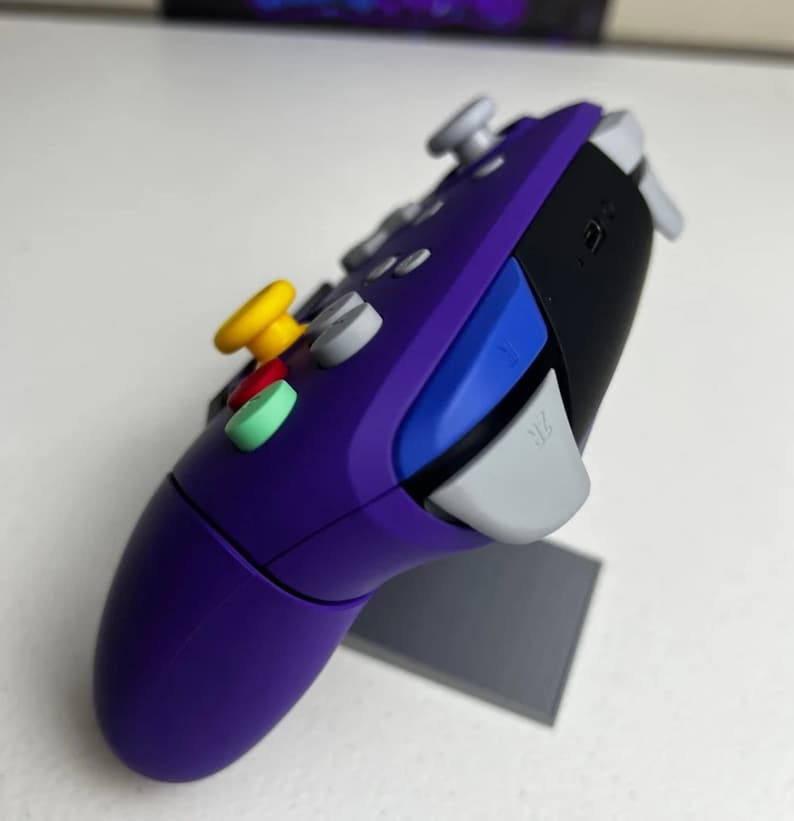 May include: Purple and black video game controller with a yellow, red, and green button. The controller has a gray button labeled "ZL".