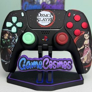 Puede incluir: Mando negro de PlayStation con diseño de "Demon Slayer". El mando tiene botones verdes, rojos y negros, y arte de personajes. El mando está sobre un soporte con el texto "GameCosmos".