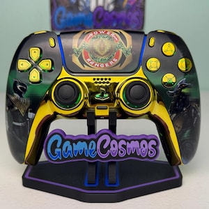 May include: A custom black and gold video game controller featuring Power Rangers graphics on the touchpad and side grips. The controller has gold buttons, black analog sticks, and is displayed on a black stand with the text GameCosmos.