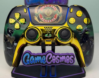 Puede incluir: Un mando de videojuegos personalizado en negro y dorado con gráficos de Power Rangers en el panel táctil y los agarres laterales. El mando tiene botones dorados, joysticks negros y se muestra en un soporte negro con el texto GameCosmos.
