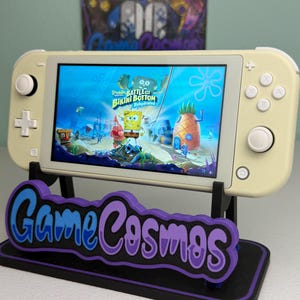 Puede incluir: Una consola de juegos portátil de color crema que muestra un juego de Bob Esponja, apoyada en un soporte negro con el texto "Game Cosmos" en azul y morado. La pantalla del juego muestra a Bob Esponja y Patricio.