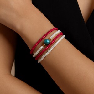 May include: A set of three beaded bracelets in red and white, with a teal bead accent. The bracelets are worn on the wrist. The beads appear to be small and uniform in size. The bracelets are a simple and elegant accessory.