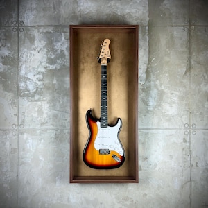Pode incluir: Uma guitarra elétrica com acabamento sunburst, exibida numa caixa de sombra de madeira. A guitarra apresenta um escudo branco, uma escala preta e cravelhas na cabeça. A caixa de sombra retangular está montada numa parede cinzenta texturizada.