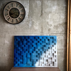 Peut inclure: Une œuvre d'art murale bleue et blanche composée de carreaux carrés. Les carreaux sont disposés en dégradé, avec les carreaux bleus à gauche et les carreaux blancs à droite. L'œuvre d'art murale est accrochée à un mur gris.