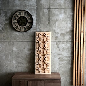 Könnte beinhalten: Eine hellbraune Akustikplatte mit geometrischem Muster hängt an einer grauen Betonwand. Die Platte ist an einem dunklen Holzkasten mit Schubladen befestigt. Eine Uhr mit römischen Ziffern und Zahnrädern befindet sich an der Wand über dem Kasten.