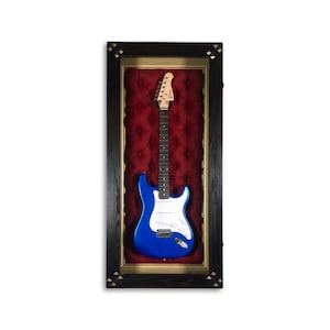 May include: A blue electric guitar displayed in a black wood frame with a red velvet backing. The frame has gold accents and decorative diamond-shaped studs.
