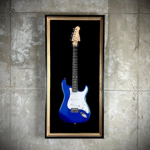 Może przedstawiać: Niebieska gitara elektryczna z białym pickguadem jest prezentowana w czarnej ramie ze złotą ramką. Gitara jest zamontowana na ścianie.