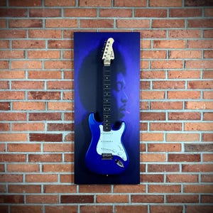 May include: A blue electric guitar with a white pickguard is mounted on a blue canvas with a silhouette of a person. The guitar is displayed against a red brick wall. The guitar's headstock has the brand name.