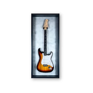 Può includere: Una chitarra elettrica con finitura sunburst, esposta in una cornice nera. La chitarra presenta una paletta di colore chiaro, una tastiera nera con intarsi a punti bianchi e un battipenna bianco. Il corpo passa dal marrone al giallo.