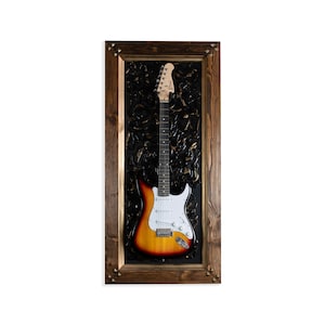 May include: A vintage-style electric guitar in a wooden frame with a black background. The guitar has a sunburst finish and a white pickguard.