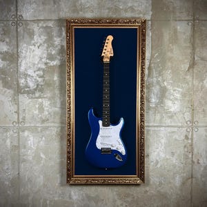 Pode incluir: Uma guitarra elétrica azul com detalhes brancos é exibida em uma moldura dourada com fundo de veludo azul.
