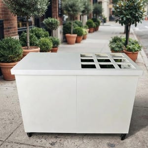 May include: A rectangular white mobile cart with a smooth top surface featuring a grid of nine square cutouts. The cart is constructed with solid white side panels and is supported by small black wheels at the base, designed for portability.