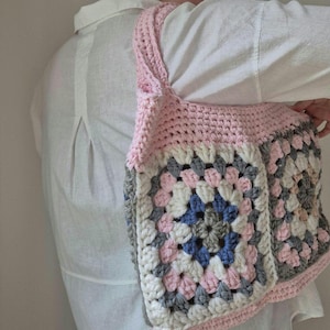 May include: A handmade crochet shoulder bag in a granny square pattern. The bag features a pink strap and trim, with a central square of blue, gray, and white. The bag is a soft, textured accessory.