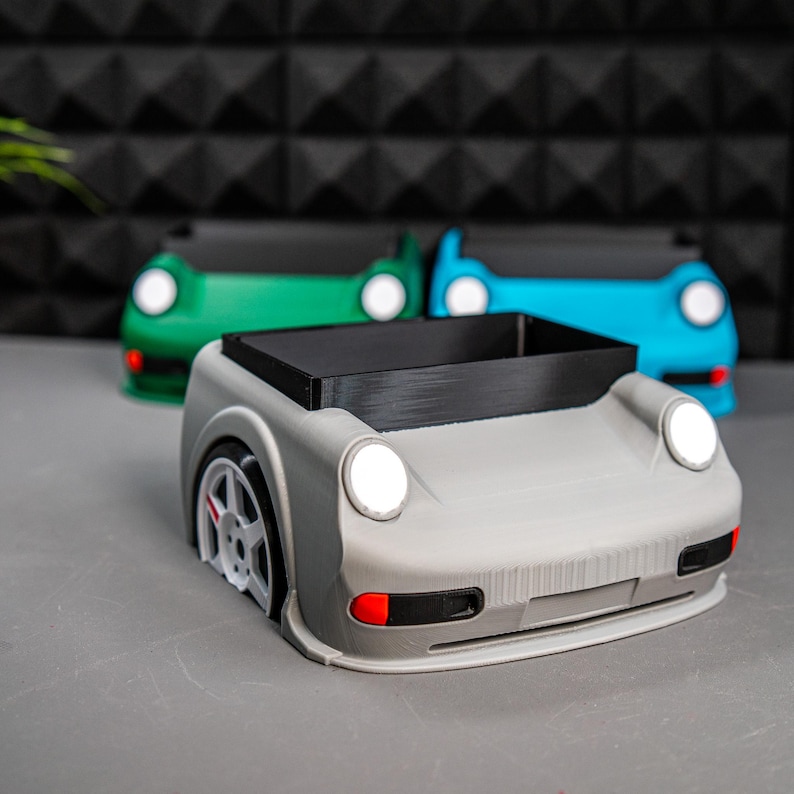 May include: Three car-shaped desk organizers in gray, green, and blue. The gray organizer has a black tray and a red accent on the front bumper.