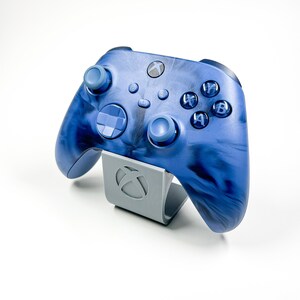 Peut inclure: Une manette Xbox bleue avec un motif en spirale sur la surface. La manette est sur un support gris avec le logo Xbox.