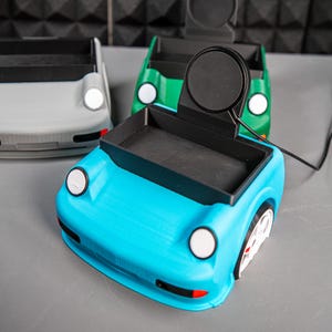 May include: A blue car-shaped phone stand with a black charging pad on top. The stand has a small tray in the back for holding small items.