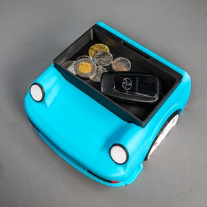 May include: A blue car-shaped trinket with a black tray inside. The tray holds a car key with the word "COROLLA" on it and several Canadian coins.