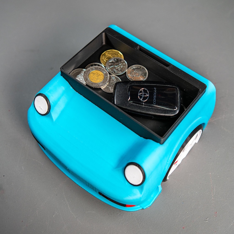May include: A blue car-shaped trinket with a black tray inside. The tray holds a car key with the word "COROLLA" on it and several Canadian coins.