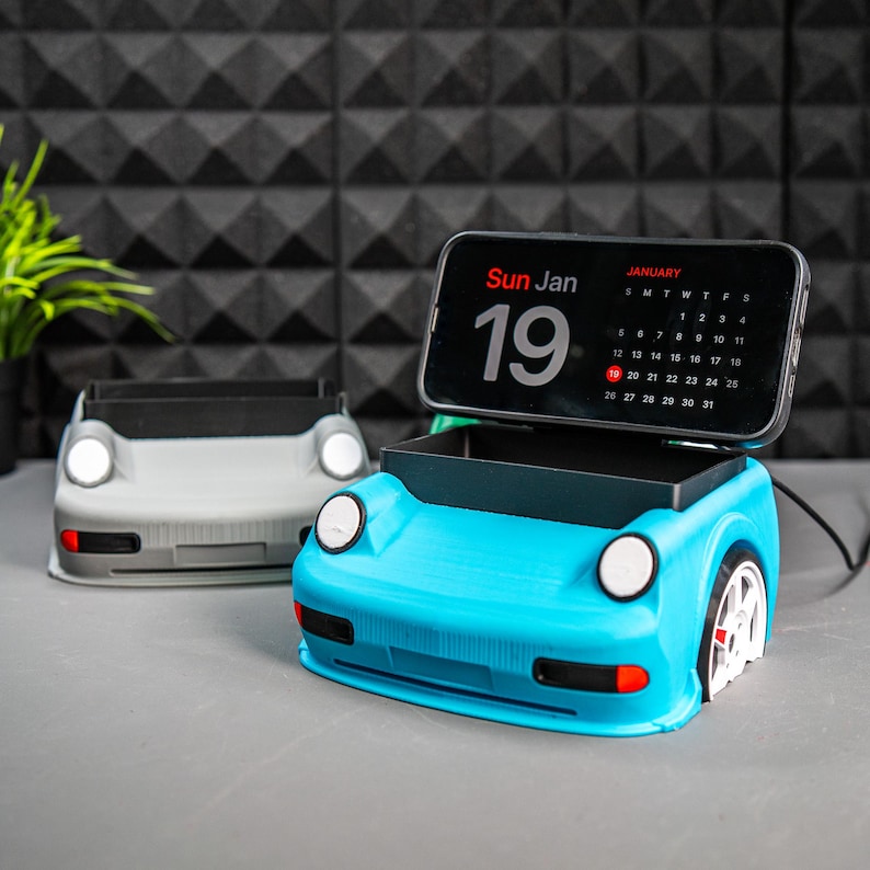 May include: A blue and gray phone stand shaped like a car. The stand has a slot for a phone and a small compartment for charging cables. The phone stand has a calendar on the phone screen that says "Sun Jan 19".