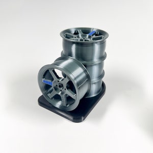 May include: A set of four gray 3D printed car wheels with blue accents. The wheels are stacked on top of each other and are sitting on a black base.