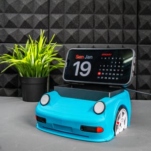 May include: A blue 3D printed phone stand shaped like a car. The stand has a slot for a phone and a calendar app is displayed on the phone screen. The date is Sunday, January 19th.