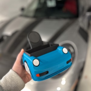 May include: A blue toy car with black accents and white headlights. The car is being held in a person's hand.